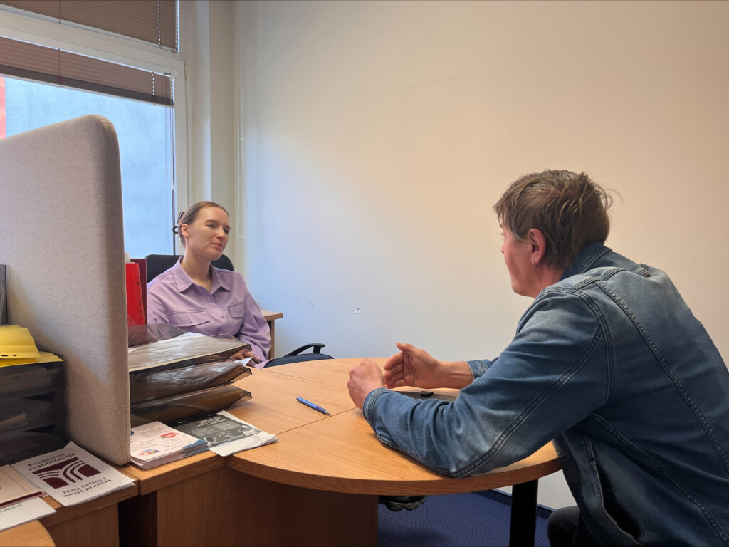 Kabinete prie darbo stalo vyksta individualus susitikimas. Moteris sėdi už stalo, o priešais ją sėdintis vyras klausosi. Ant stalo matyti dokumentų dėklai, lankstinukai ir rašiklis. Susitikimas vyksta ramioje darbo aplinkoje, apšviestoje dienos šviesos iš lango.