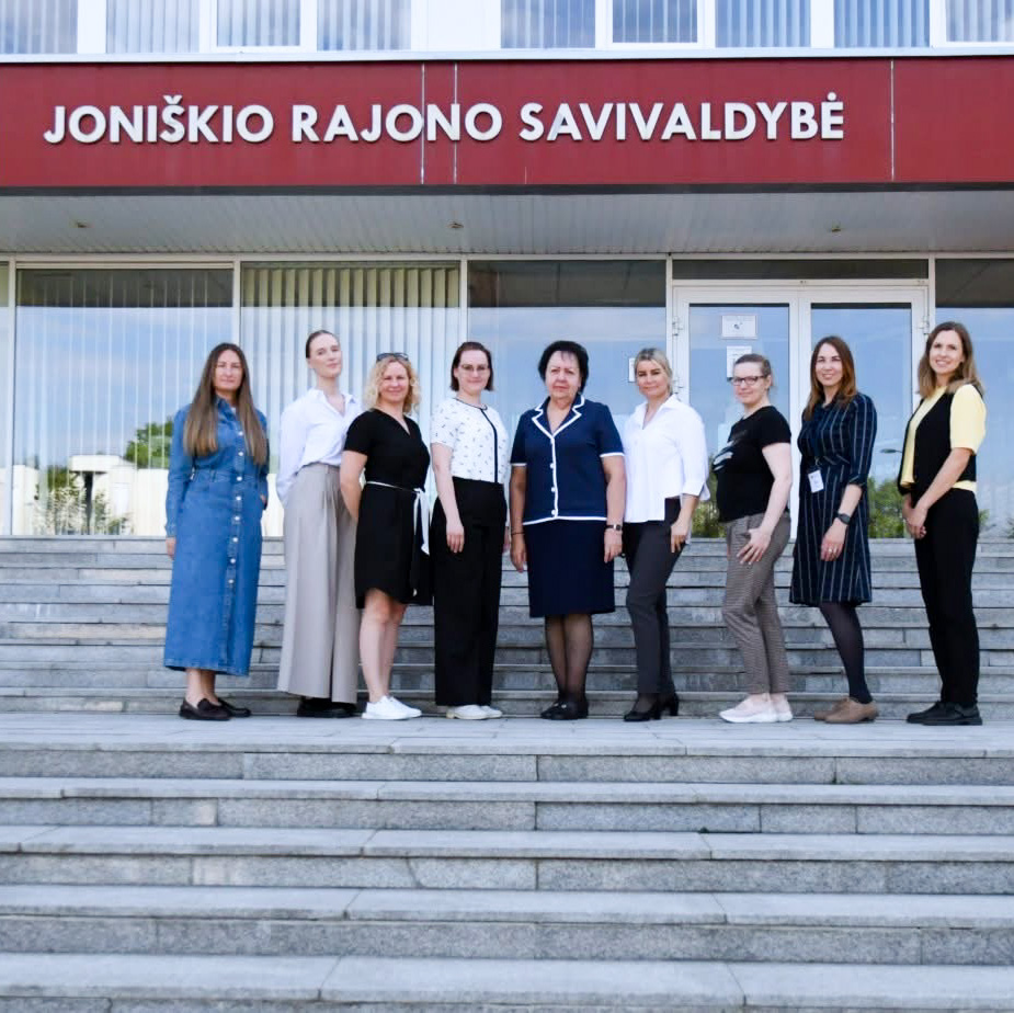 Prie Joniškio rajono savivaldybės pastato pagrindinio įėjimo ant laiptų stovi devynios moterys – Šiaulių regiono atvejo vadybininkės ir Joniškio rajono savivaldybės atstovės. Virš jų matomas užrašas „Joniškio rajono savivaldybė“. Dalyvės pozuoja bendrai nuotraukai lauke, priešais savivaldybės pastatą.