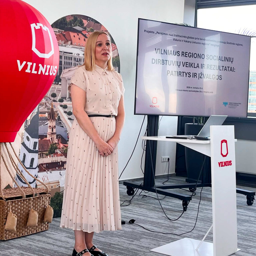 Šviesioje konferencijų salėje moteris stovi priekyje ir kalba renginio dalyviams. Ji vilki šviesią, kreminės spalvos suknelę su smulkiais taškeliais ir juodu dirželiu. Šalia jos stovi ekranas, kuriame rodoma prezentacija apie Vilniaus regiono socialinių dirbtuvių veiklą ir rezultatus. Kairėje matoma dekoratyvinė raudona oro baliono instaliacija su Vilniaus miesto logotipu, fone – miesto panoramos stendas. Dešinėje – tribūna su Vilniaus logotipu ir stalas su nešiojamuoju kompiuteriu. Salė moderni, gerai apšviesta, pro langus patenka dienos šviesa.