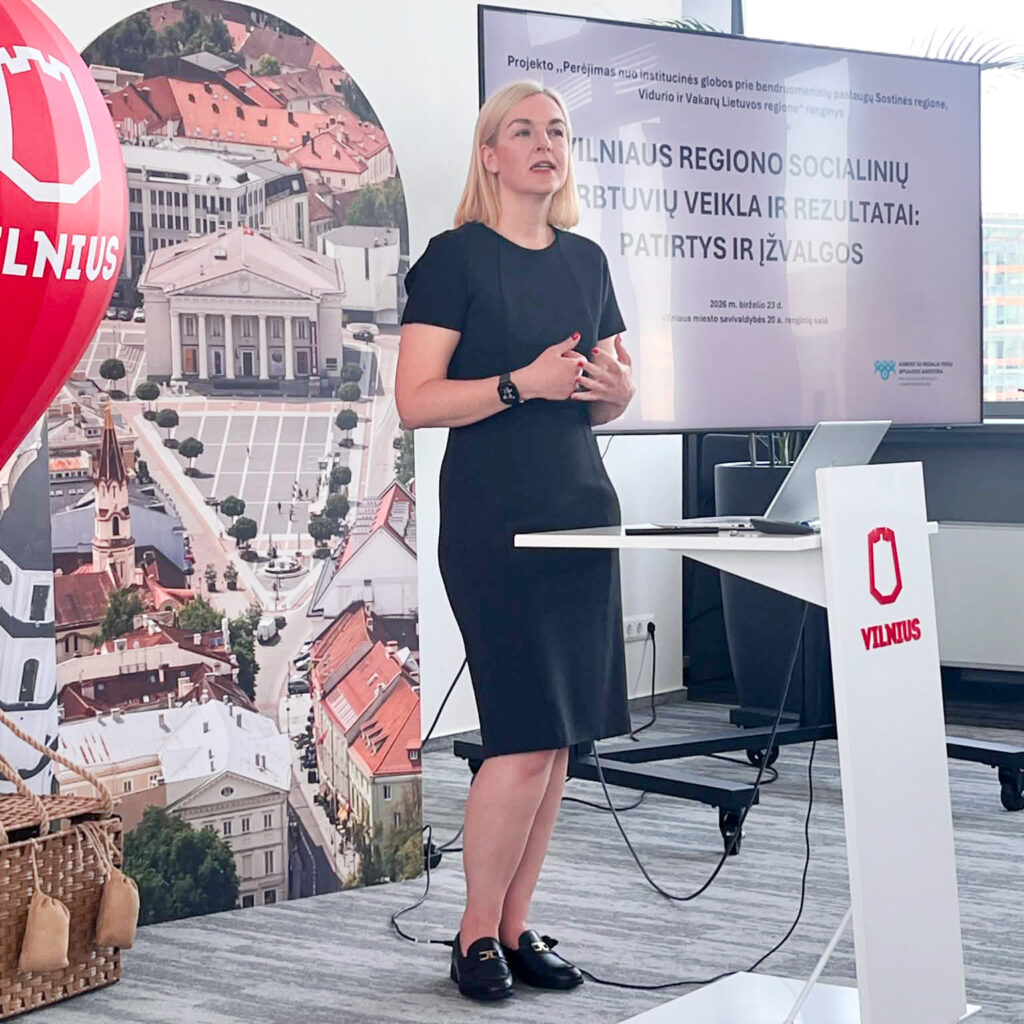 Konferencijų salėje moteris juoda suknele stovi priekyje ir skaito pranešimą, kalbėdama gestikuliuoja rankomis. Už jos esančiame ekrane rodoma pristatymo skaidrė „Vilniaus regiono socialinių dirbtuvių veikla ir rezultatai: patirtys ir įžvalgos“. Kairėje matoma dekoratyvinė raudona oro baliono instaliacija su Vilniaus miesto logotipu ir miesto panoramos stendas. Dešinėje – tribūna su Vilniaus logotipu, stalas su nešiojamuoju kompiuteriu ir prezentacijos įranga. Moderni salė apšviesta natūralia dienos šviesa, sklindančia pro didelius langus.