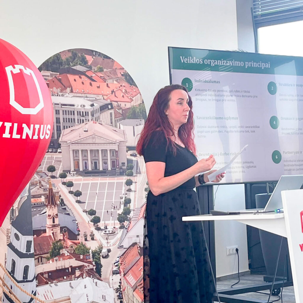 Konferencijų salėje moteris tamsia suknele stovi priekyje ir skaito pranešimą, vienoje rankoje laikydama užrašus, kita gestikuliuodama. Už jos esančiame ekrane rodoma skaidrė „Veiklos organizavimo principai“, kurioje pateikti pagrindiniai socialinių dirbtuvių veiklos principai. Kairėje matoma dekoratyvinė raudona oro baliono instaliacija su Vilniaus miesto logotipu ir miesto panoramos stendas. Dešinėje – tribūna su Vilniaus logotipu, stalas su nešiojamuoju kompiuteriu ir prezentacijos įranga. Moderni salė apšviesta natūralia dienos šviesa, sklindančia pro didelius langus.