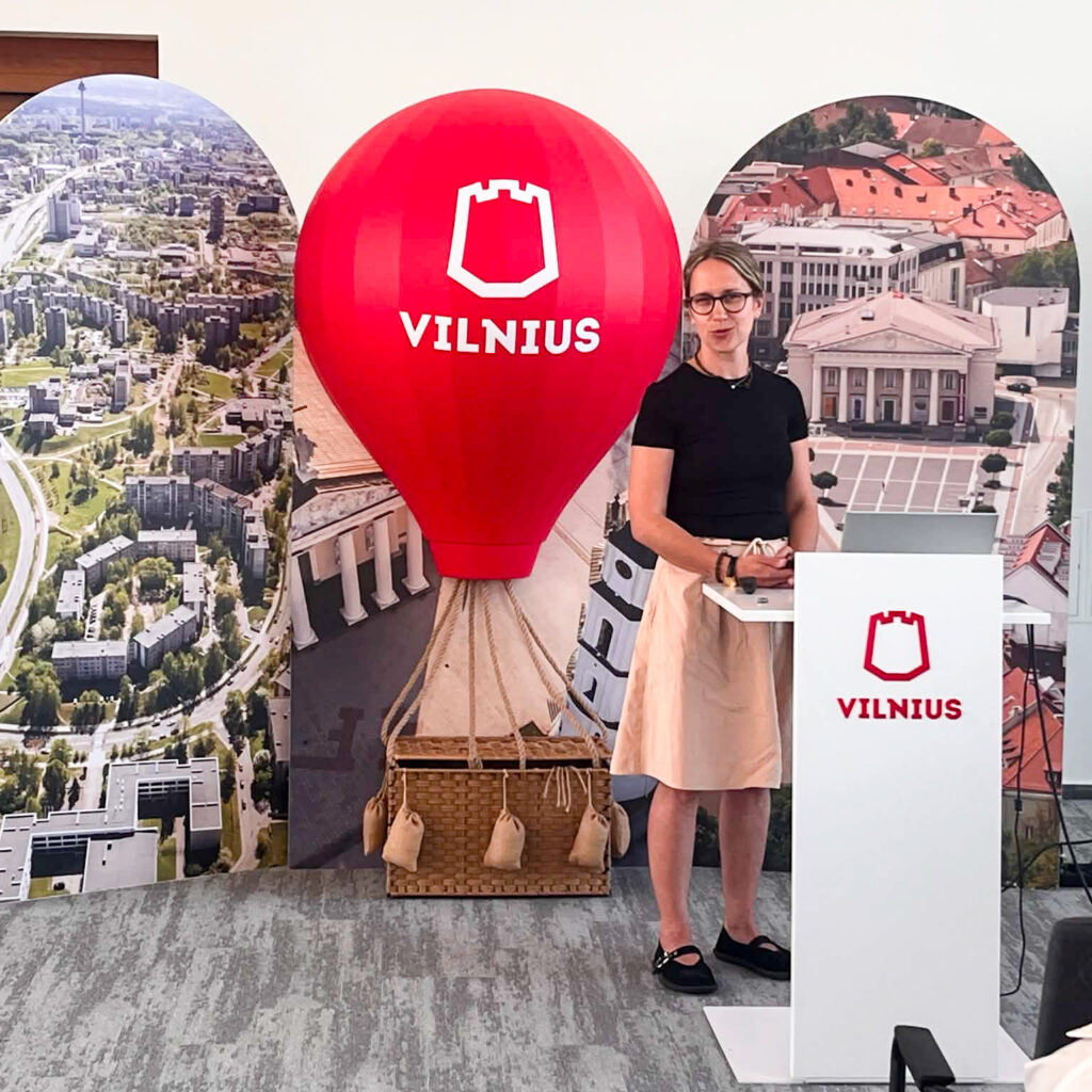 Konferencijų salėje moteris stovi už baltos tribūnos su Vilniaus miesto logotipu ir skaito pranešimą. Ji vilki juodus marškinėlius, šviesų sijoną, avi juodus batelius ir dėvi akinius. Už jos matoma didelė dekoratyvinė raudona oro baliono instaliacija su Vilniaus miesto logotipu, o iš abiejų pusių – Vilniaus panoramos stendai. Ant tribūnos padėtas nešiojamasis kompiuteris ir prezentacijos valdymo įranga. Moderni konferencijų salė erdvi ir šviesi.