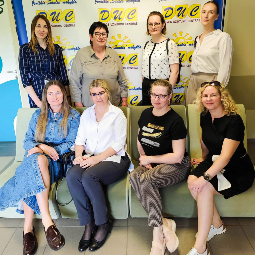 Grupinė nuotrauka Joniškio „Saulės“ mokyklos Dienos užimtumo centre. Prie centro stendo su įstaigos logotipais pozuoja aštuonios moterys – Šiaulių regiono atvejo vadybininkės ir centro atstovės. Keturios moterys stovi, keturios sėdi ant minkštasuolio. Nuotrauka daryta centro patalpose vizito metu.