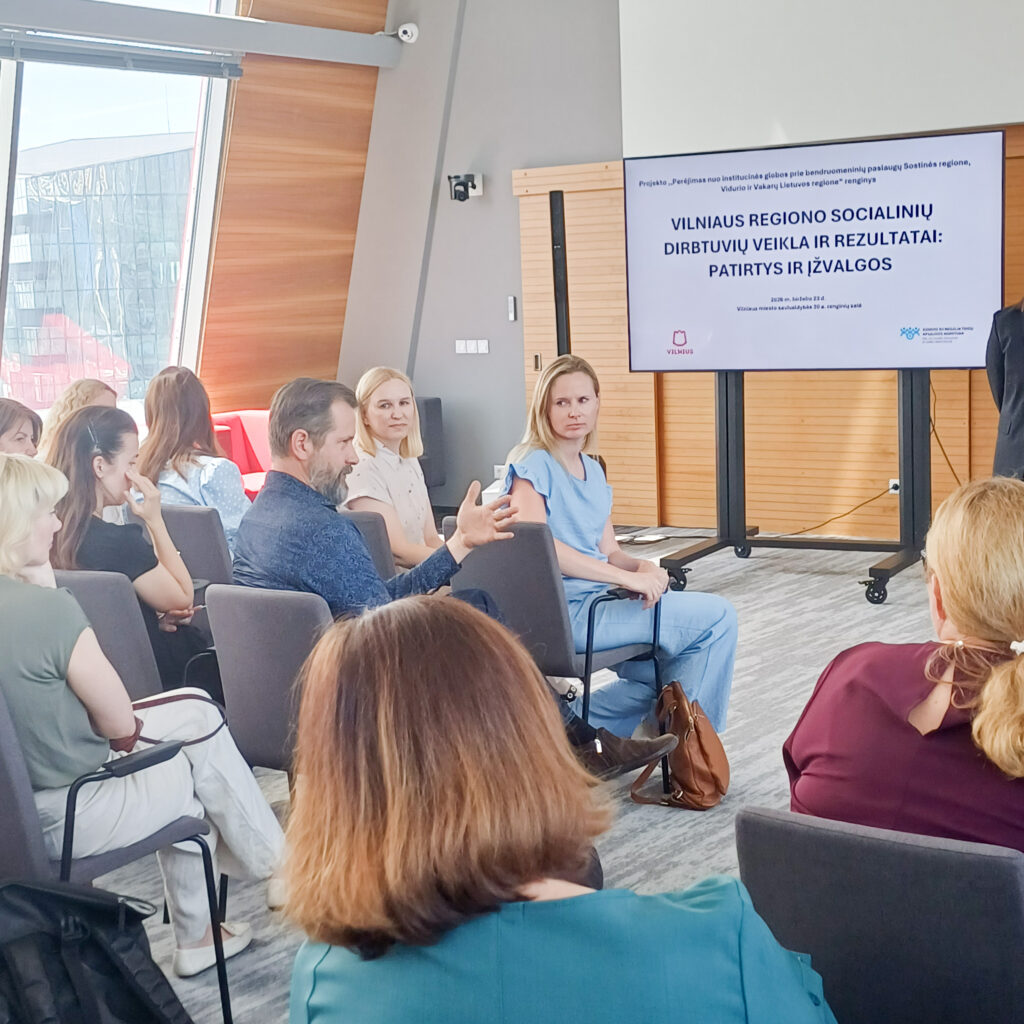 Konferencijų salėje vyksta diskusija po pranešimo. Pirmose eilėse sėdintis vyras kalba ir gestikuliuoja ranka, o kiti dalyviai klausosi bei stebi jį. Fone matomas ekranas su pristatymo skaidre „Vilniaus regiono socialinių dirbtuvių veikla ir rezultatai: patirtys ir įžvalgos“. Salėje sėdi keliolika renginio dalyvių, išdėstytų kėdėse puslankiu. Erdvi moderni salė apšviesta natūralia dienos šviesa, sklindančia pro didelius langus.