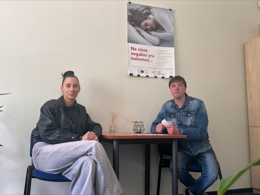 Kabinete prie nedidelio stalo sėdi moteris ir vyras. Abu atsisukę į kamerą. Ant stalo yra dekoratyvi žvakidė ir nedidelė vazelė su džiovintais augalais. Ant sienos virš stalo kabo plakatas, skirtas atkreipti dėmesį į nematomas negalias. Nuotrauka daryta ramioje darbo ar konsultacijų aplinkoje.