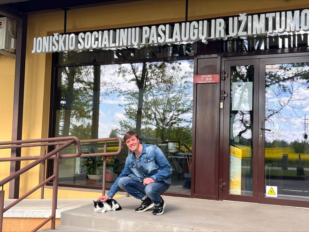 Lauke prie Joniškio socialinių paslaugų ir užimtumo centro įėjimo vyras pritūpęs šalia ant laiptelio gulinčio juodai balto katino ir jį glosto. Už jų matyti pastato stiklinės durys, dideli langai ir virš įėjimo esantis įstaigos pavadinimas. Languose atsispindi medžiai ir aplinka.