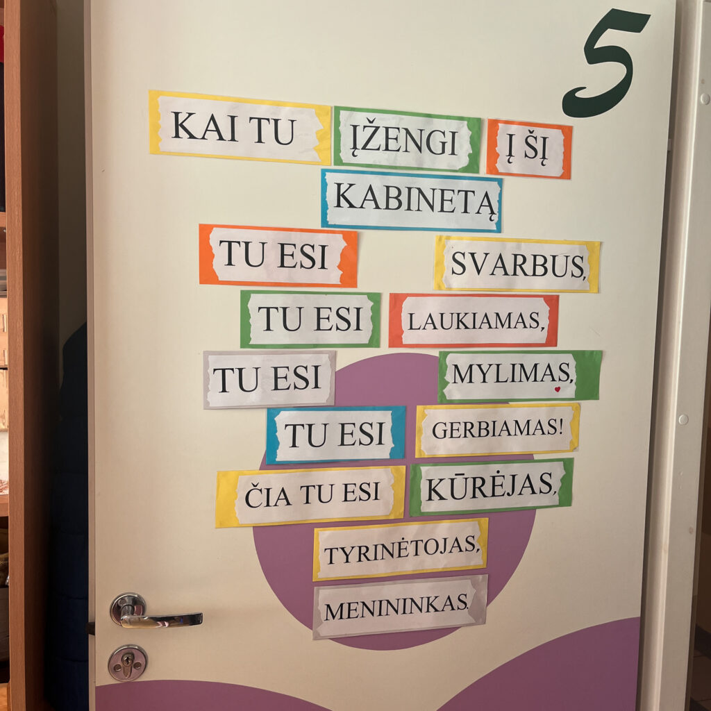Ant baltų durų pritvirtintas motyvuojantis užrašas iš spalvotų kortelių. Jame rašoma: „Kai tu įžengi į šį kabinetą, tu esi svarbus, tu esi laukiamas, tu esi mylimas, tu esi gerbiamas! Čia tu esi kūrėjas, tyrinėtojas, menininkas.“ Durų apačia dekoruota šviesiai violetiniais grafiniais elementais.