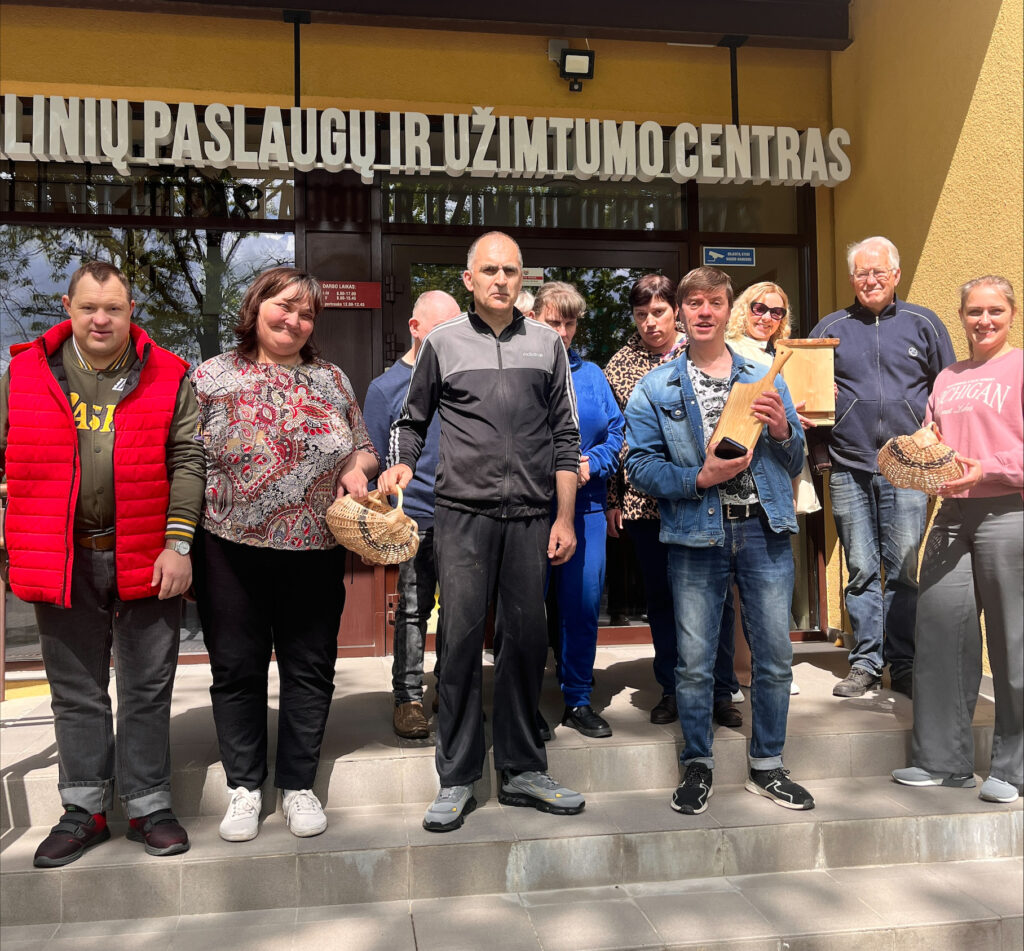 Prie Joniškio socialinių paslaugų ir užimtumo centro įėjimo susibūrusi grupė žmonių pozuoja bendrai nuotraukai. Kai kurie dalyviai rankose laiko savo pagamintus dirbinius – pintus krepšelius ir medinius gaminius. Grupė stovi ant laiptų prie pagrindinio įėjimo, virš kurio matyti įstaigos pavadinimas.