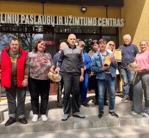 Prie Joniškio socialinių paslaugų ir užimtumo centro įėjimo susibūrusi grupė žmonių pozuoja bendrai nuotraukai. Kai kurie dalyviai rankose laiko savo pagamintus dirbinius – pintus krepšelius ir medinius gaminius. Grupė stovi ant laiptų prie pagrindinio įėjimo, virš kurio matyti įstaigos pavadinimas.