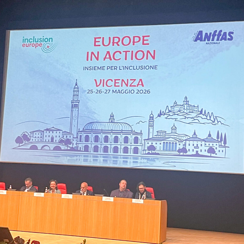 Konferencijos „Europe in Action 2026“ atidarymo sesija Vičencoje. Prie ilgo stalo scenoje sėdi keli pranešėjai, o virš jų dideliame ekrane rodomas renginio pavadinimas, organizatorių logotipai ir konferencijos datos. Salėje vyksta oficialus renginio pristatymas.