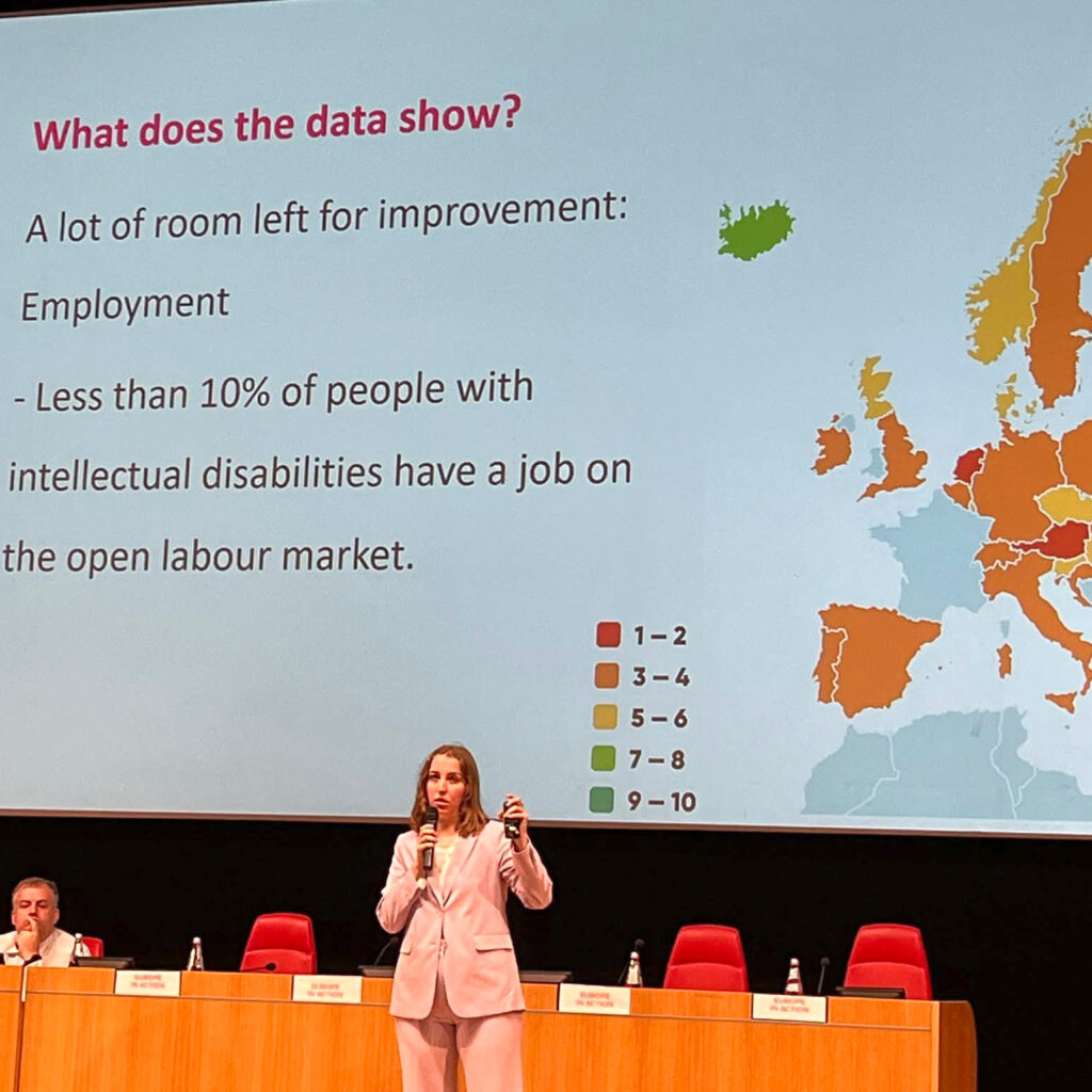 Konferencijoje pranešėja stovi scenoje ir kalba auditorijai, rankoje laikydama mikrofoną. Už jos dideliame ekrane rodoma skaidrė apie žmonių su intelekto negalia užimtumą Europoje. Skaidrėje pateikiamas Europos žemėlapis ir duomenys, rodantys, kad mažiau nei 10 proc. žmonių su intelekto negalia dirba atviroje darbo rinkoje.