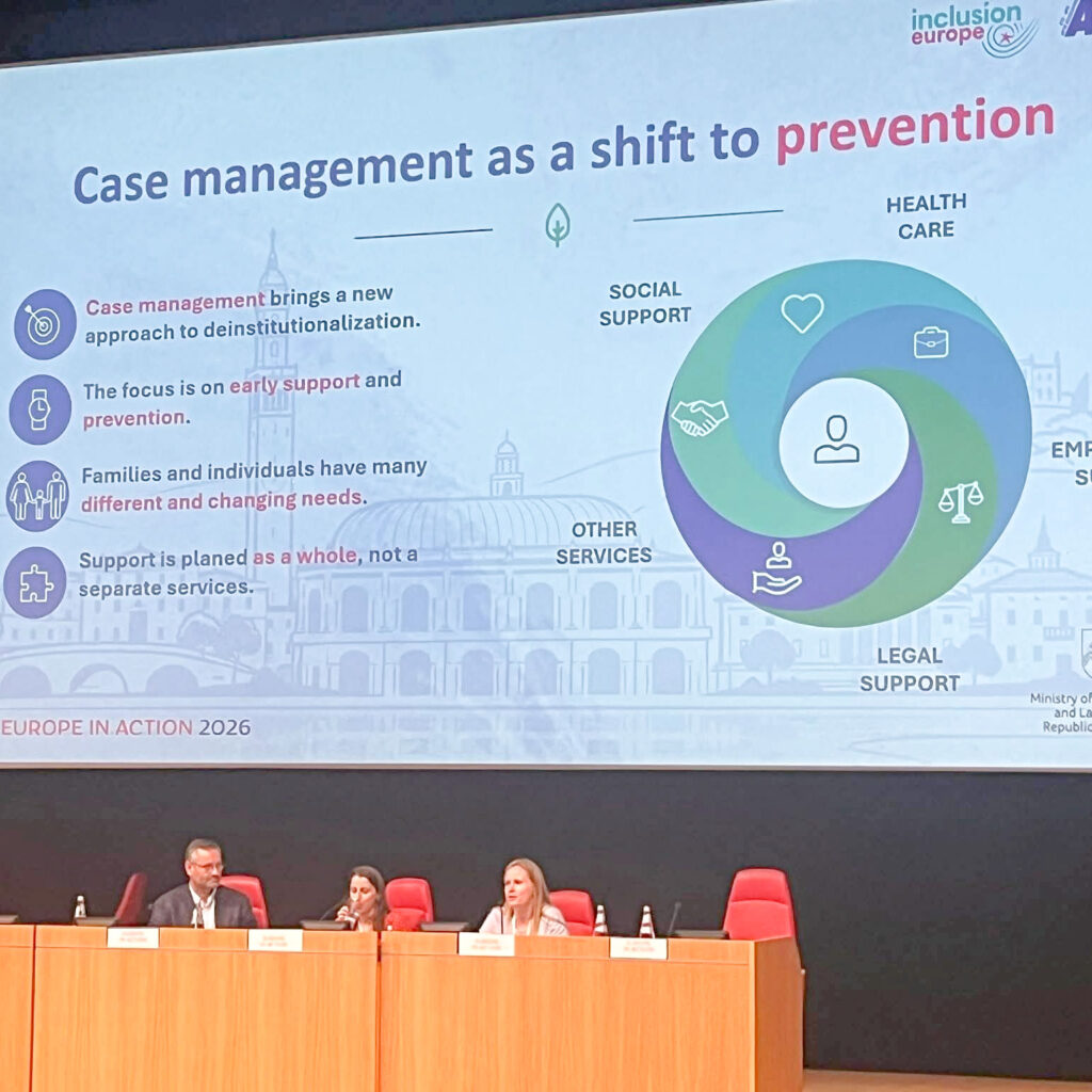Konferencijos salėje vyksta pranešimas apie atvejo vadybą kaip prevencijos priemonę. Dideliame ekrane rodoma skaidrė, kurioje aiškinama, kaip socialinė, sveikatos priežiūros, teisinė ir kita pagalba gali būti koordinuojama siekiant užkirsti kelią institucinės globos poreikiui. Priekyje prie stalo sėdi trys pranešėjai.
