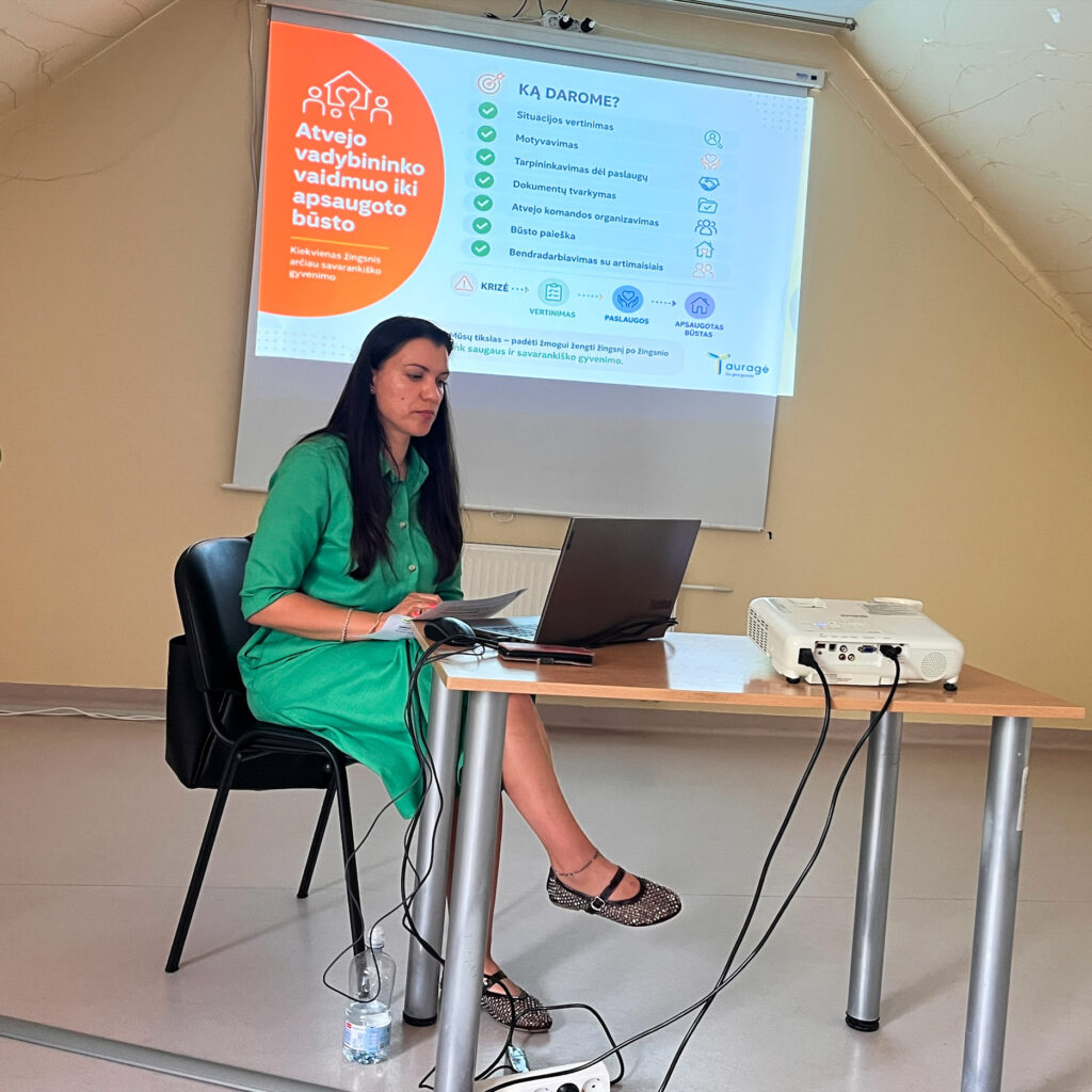 Moteris sėdi prie stalo ir skaito pranešimą, naudodamasi nešiojamuoju kompiuteriu. Už jos ekrane rodoma prezentacija apie atvejo vadybininko vaidmenį organizuojant apsaugoto būsto paslaugą ir pagalbą žmogui siekiant savarankiško gyvenimo. Ant stalo matyti projektorius, o šalia – vandens buteliukas.