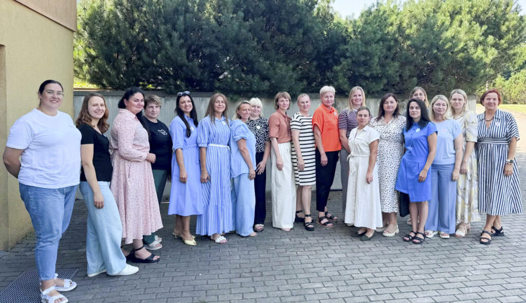 Grupinė Marijampolės ir Tauragės regionų specialistų nuotrauka. Devyniolika moterų stovi lauke prie pastato ir žvelgia į fotoaparatą. Už jų – žaliuojantys medžiai ir krūmai.