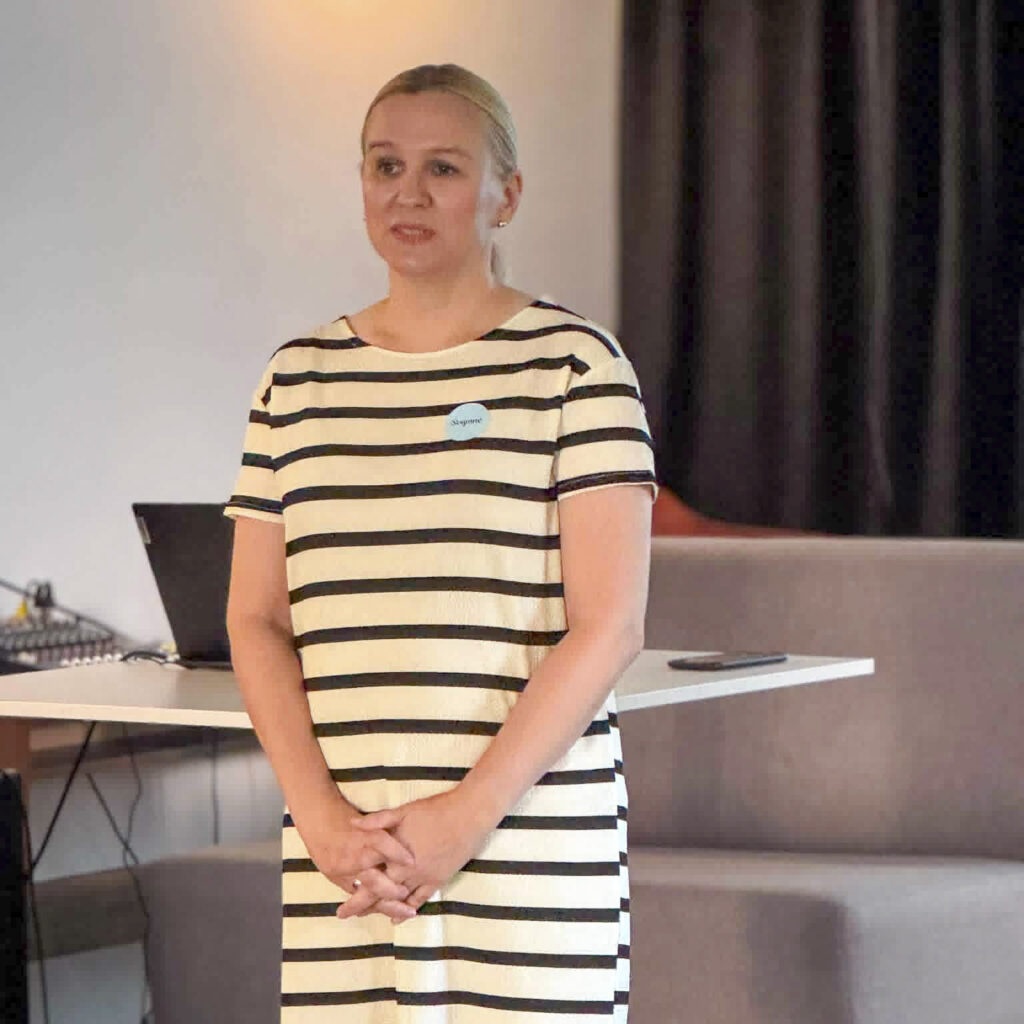 Projekto vadovė Svajonė Rainienė stovi salėje ir kalba susitikimo dalyviams. Už jos matomas stalas su nešiojamuoju kompiuteriu ir sėdimos vietos.
