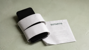 Šviesiai žaliame fone guli juodas išmanusis telefonas, apjuostas baltomis popieriaus juostomis. Šalia – dokumentas su užrašu „SUTARTIS“.